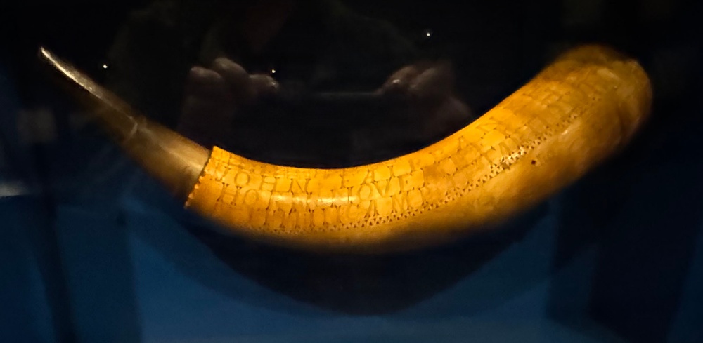 The image shows an engraved animal horn, likely a powder horn or drinking horn, displayed in what appears to be a museum setting under dramatic lighting. The horn is curved and yellowish-amber in color with visible inscribed text and decorative dot patterns along its surface. The name 'JOHN' and partial additional text are legible among the engravings.