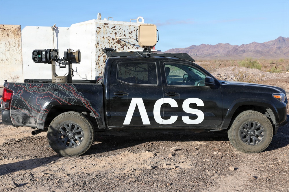 A black Toyota Tacoma pickup truck branded with 'ACS' lettering is parked in a desert environment, likely a test range in the American Southwest. The truck has been modified with specialized equipment mounted in the bed, including what appears to be a sensor/camera gimbal system and a radar or antenna unit on the roof. The vehicle features topographic map-style graphics on the lower body panels.