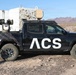 A black Toyota Tacoma pickup truck branded with 'ACS' lettering is parked in a desert environment, likely a test range in the American Southwest. The truck has been modified with specialized equipment mounted in the bed, including what appears to be a sensor/camera gimbal system and a radar or antenna unit on the roof. The vehicle features topographic map-style graphics on the lower body panels.