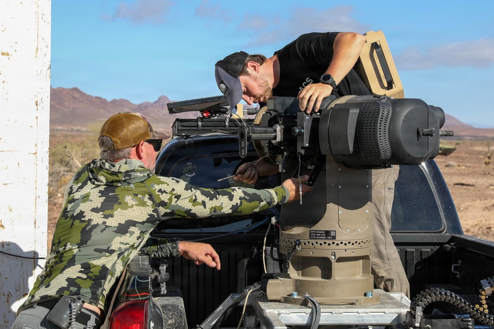 Two individuals are working on a mounted weapon or sensor system installed in the bed of a pickup truck in a desert environment. One person wearing camouflage clothing is working from the side while another in a black shirt leans over the equipment from above. The setting appears to be an outdoor testing or demonstration range based on the arid landscape and makeshift structure visible.