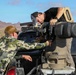 Two individuals are working on a mounted weapon or sensor system installed in the bed of a pickup truck in a desert environment. One person wearing camouflage clothing is working from the side while another in a black shirt leans over the equipment from above. The setting appears to be an outdoor testing or demonstration range based on the arid landscape and makeshift structure visible.