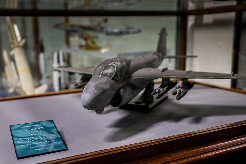 A museum display case features a scale model of a Grumman A-6A Intruder strike aircraft, mounted on a gray surface. The accompanying placard identifies it as recovered from the Pentagon Building following the crash of American Airlines Flight 77 on September 11, 2001. The model belongs to the Department of the Navy and bears catalog number 2130.