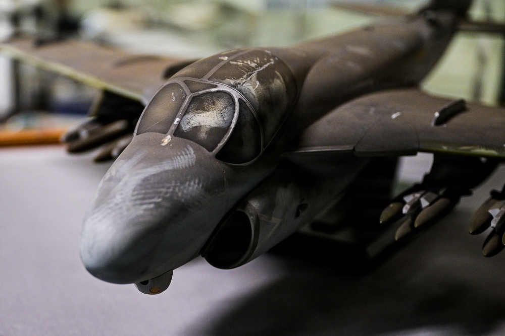 The image shows a detailed scale model of a military jet aircraft, likely an AV-8B Harrier II or similar ground-attack aircraft, placed on a flat surface. The model features dark gray/olive paint scheme with visible weathering effects, a multi-panel cockpit canopy, and multiple ordnance/weapons mounted under the wings. The close-up perspective emphasizes the nose and forward fuselage section with fine surface detailing.