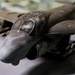 The image shows a detailed scale model of a military jet aircraft, likely an AV-8B Harrier II or similar ground-attack aircraft, placed on a flat surface. The model features dark gray/olive paint scheme with visible weathering effects, a multi-panel cockpit canopy, and multiple ordnance/weapons mounted under the wings. The close-up perspective emphasizes the nose and forward fuselage section with fine surface detailing.