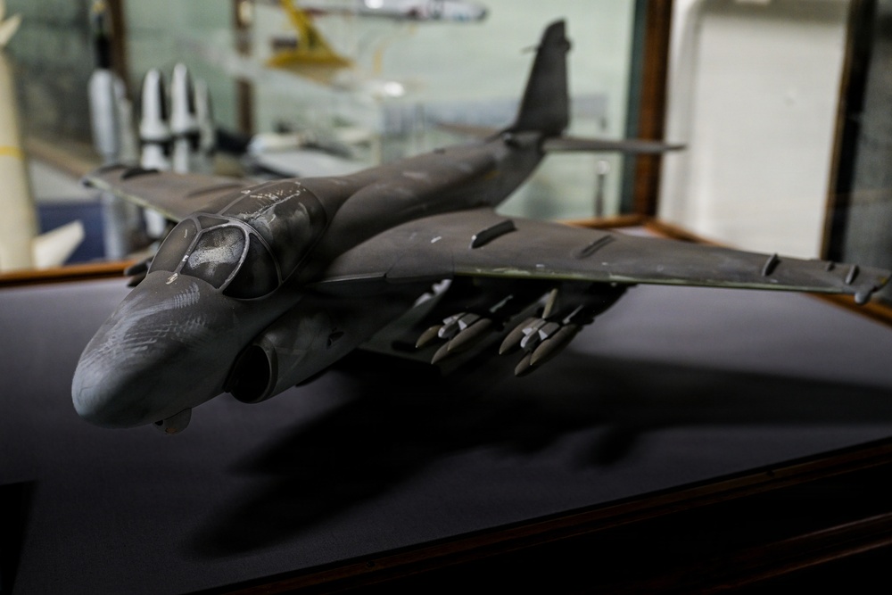 The image shows a detailed scale model of a military jet aircraft, likely a B-1 Lancer or similar swept-wing bomber, displayed on a dark surface inside what appears to be a museum or display case setting. The model is painted in a dark gray/charcoal color scheme and features multiple munitions mounted under the fuselage. Additional aircraft models and display cases are visible in the blurred background.