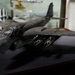 The image shows a detailed scale model of a military jet aircraft, likely a B-1 Lancer or similar swept-wing bomber, displayed on a dark surface inside what appears to be a museum or display case setting. The model is painted in a dark gray/charcoal color scheme and features multiple munitions mounted under the fuselage. Additional aircraft models and display cases are visible in the blurred background.