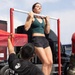 A young woman in athletic wear performs a pull-up on a red portable pull-up bar while a U.S. Marine in camouflage pants and a black USMC t-shirt stabilizes the equipment. The setting appears to be an outdoor public event or recruitment demonstration, with large trucks and a red tent visible in the background. The Marine's shirt reads 'Pain is Weakness Leaving the Body,' a well-known Marine Corps motivational phrase.