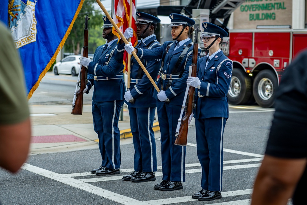 A United States Air Force Honor Guard color guard team stands at attention on a street during a formal ceremony. Four service members in dress blue uniforms are positioned in formation, with two carrying flags and two carrying ceremonial rifles. The setting appears to be an outdoor public event near a governmental building.