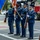 A United States Air Force Honor Guard color guard team stands at attention on a street during a formal ceremony. Four service members in dress blue uniforms are positioned in formation, with two carrying flags and two carrying ceremonial rifles. The setting appears to be an outdoor public event near a governmental building.