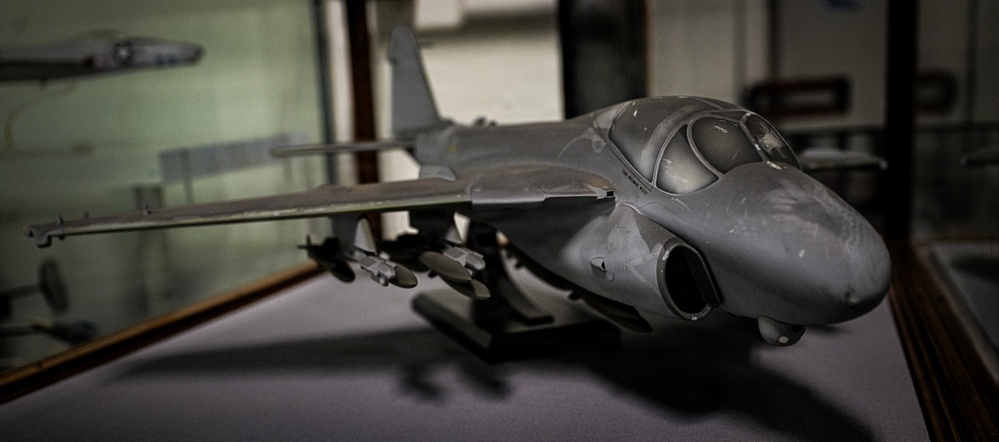 The image shows a detailed scale model of a military jet aircraft, painted in overall gray, displayed on a flat surface. The model features underwing ordnance, landing gear deployed, and a prominent nose with a two-seat tandem cockpit configuration, consistent with an aircraft such as the Grumman EA-6B Prowler or A-6 Intruder. The setting appears to be a display case or workshop environment with another aircraft model visible in the blurred background.