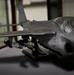The image shows a detailed scale model of a military jet aircraft, painted in overall gray, displayed on a flat surface. The model features underwing ordnance, landing gear deployed, and a prominent nose with a two-seat tandem cockpit configuration, consistent with an aircraft such as the Grumman EA-6B Prowler or A-6 Intruder. The setting appears to be a display case or workshop environment with another aircraft model visible in the blurred background.
