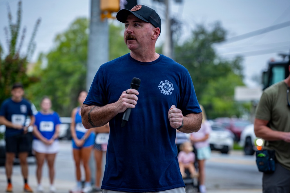 A man wearing a navy blue Fire Rescue t-shirt and black cap is speaking into a microphone at an outdoor public event. He appears to be addressing a crowd gathered on a street or parking area, with several people visible in the background wearing race bibs, suggesting this may be a charity run or community event. A fire truck or emergency vehicle is partially visible on the right side of the frame.