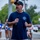 A man wearing a navy blue Fire Rescue t-shirt and black cap is speaking into a microphone at an outdoor public event. He appears to be addressing a crowd gathered on a street or parking area, with several people visible in the background wearing race bibs, suggesting this may be a charity run or community event. A fire truck or emergency vehicle is partially visible on the right side of the frame.
