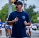 A man wearing a navy blue Fire Rescue t-shirt and black cap is speaking into a microphone at an outdoor public event. He appears to be addressing a crowd gathered on a street or parking area, with several people visible in the background wearing race bibs, suggesting this may be a charity run or community event. A fire truck or emergency vehicle is partially visible on the right side of the frame.