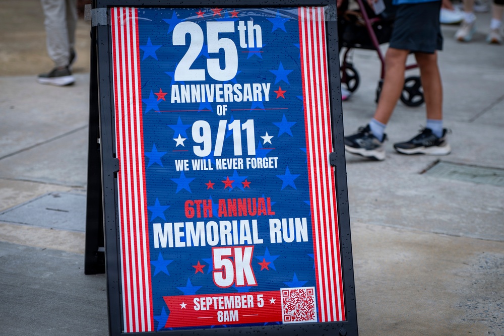 A patriotic A-frame sandwich board sign advertises the 25th Anniversary of 9/11 Memorial Run 5K event. The sign promotes the '6th Annual Memorial Run 5K' scheduled for September 5 at 8AM, with the tagline 'We Will Never Forget.' The sign is displayed on a sidewalk with pedestrians visible in the background.