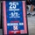 A patriotic A-frame sandwich board sign advertises the 25th Anniversary of 9/11 Memorial Run 5K event. The sign promotes the '6th Annual Memorial Run 5K' scheduled for September 5 at 8AM, with the tagline 'We Will Never Forget.' The sign is displayed on a sidewalk with pedestrians visible in the background.