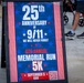A patriotic A-frame sandwich board sign advertises the 25th Anniversary of 9/11 Memorial Run 5K event. The sign promotes the '6th Annual Memorial Run 5K' scheduled for September 5 at 8AM, with the tagline 'We Will Never Forget.' The sign is displayed on a sidewalk with pedestrians visible in the background.