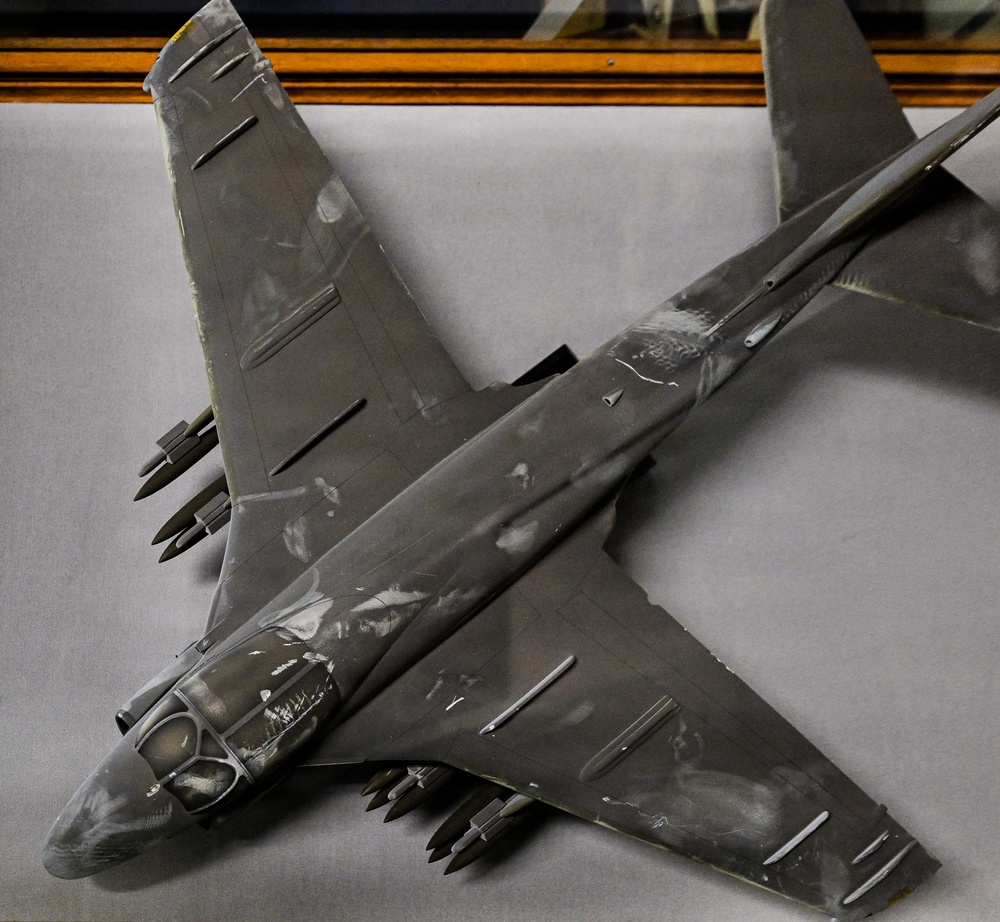 The image shows a detailed scale model of a military jet aircraft, displayed on a flat gray surface. The model appears to represent a Cold War-era bomber or attack aircraft, finished in a dark gray paint scheme and equipped with multiple underwing ordnance. A framed picture or display case is partially visible in the background.