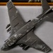 The image shows a detailed scale model of a military jet aircraft, displayed on a flat gray surface. The model appears to represent a Cold War-era bomber or attack aircraft, finished in a dark gray paint scheme and equipped with multiple underwing ordnance. A framed picture or display case is partially visible in the background.