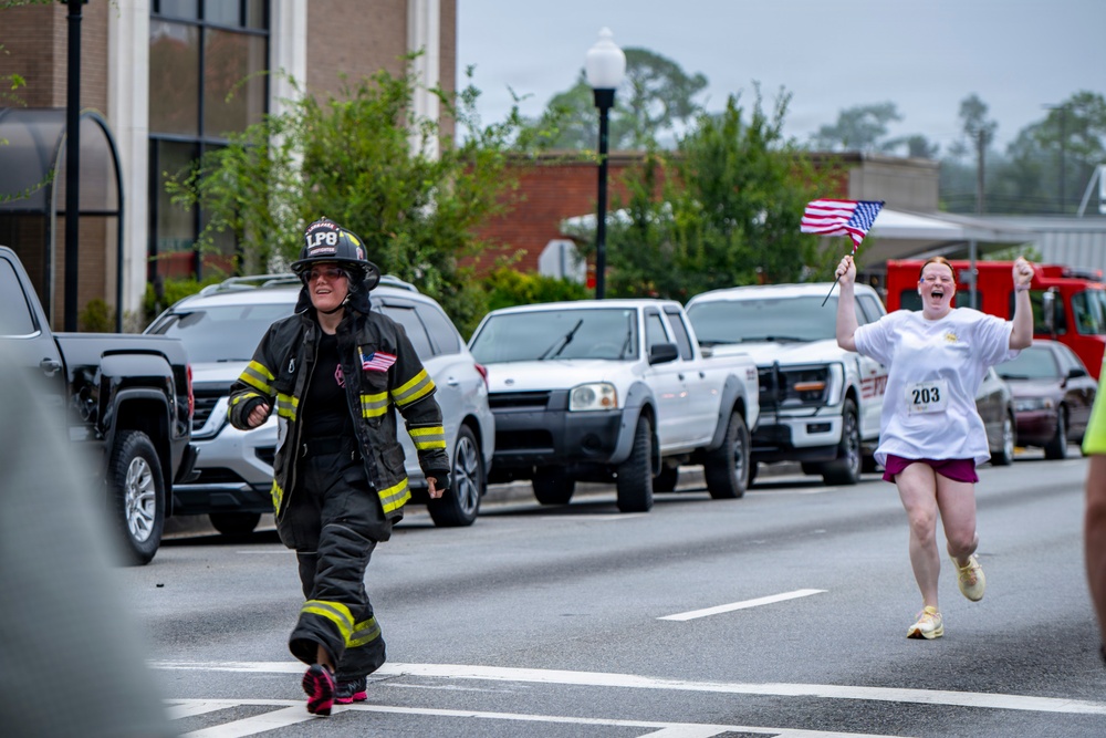 Two women are participating in a road race on a city street. One runner is dressed in full firefighter turnout gear including a helmet marked 'LP8', while the other wears a white race shirt with bib number 203 and is enthusiastically waving a small American flag. Both appear to be in high spirits, smiling and celebrating as they run.