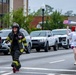 Two women are participating in a road race on a city street. One runner is dressed in full firefighter turnout gear including a helmet marked 'LP8', while the other wears a white race shirt with bib number 203 and is enthusiastically waving a small American flag. Both appear to be in high spirits, smiling and celebrating as they run.