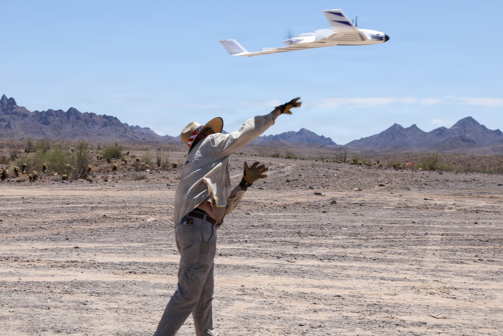 A person in a desert environment has just hand-launched a fixed-wing UAV (unmanned aerial vehicle) into the air. The aircraft, white with blue markings, is captured mid-flight just after release, with the operator's arms still extended in the throwing motion. The arid landscape features sparse desert vegetation, rocky terrain, and mountain ranges in the background under a clear blue sky.