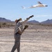 A person in a desert environment has just hand-launched a fixed-wing UAV (unmanned aerial vehicle) into the air. The aircraft, white with blue markings, is captured mid-flight just after release, with the operator's arms still extended in the throwing motion. The arid landscape features sparse desert vegetation, rocky terrain, and mountain ranges in the background under a clear blue sky.