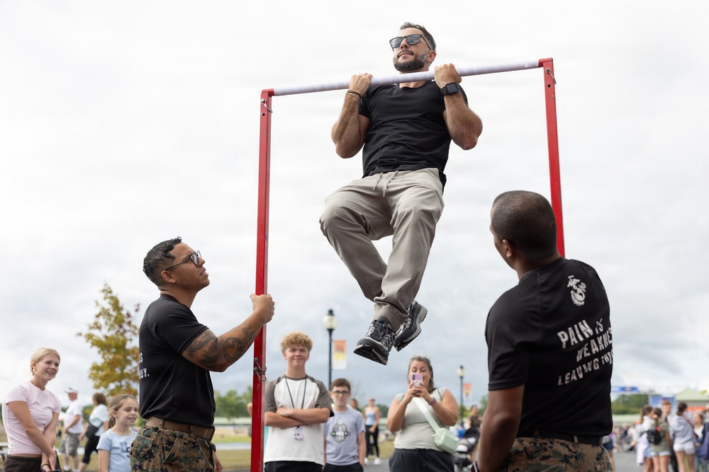 A civilian man in a black t-shirt and khaki pants is performing pull-ups on a red pull-up bar at what appears to be a public military recruitment or outreach event. Two Marines in black PT shirts and camouflage trousers flank the bar, one steadying it and one observing. A crowd of spectators, including teenagers and adults, watches in the background.