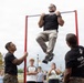A civilian man in a black t-shirt and khaki pants is performing pull-ups on a red pull-up bar at what appears to be a public military recruitment or outreach event. Two Marines in black PT shirts and camouflage trousers flank the bar, one steadying it and one observing. A crowd of spectators, including teenagers and adults, watches in the background.
