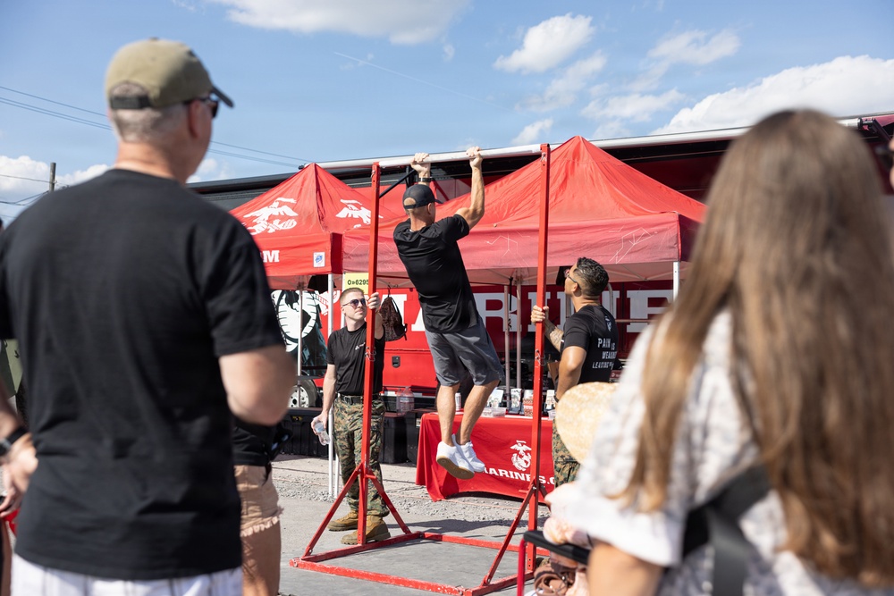A public U.S. Marines recruiting event is taking place outdoors, featuring a red pull-up bar station where a civilian in a black t-shirt and gray shorts is performing pull-ups. Two Marines in camouflage uniforms are observing and monitoring the activity. Spectators, including a man in a black shirt and a woman with brown hair, watch from the foreground.