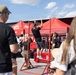 A public U.S. Marines recruiting event is taking place outdoors, featuring a red pull-up bar station where a civilian in a black t-shirt and gray shorts is performing pull-ups. Two Marines in camouflage uniforms are observing and monitoring the activity. Spectators, including a man in a black shirt and a woman with brown hair, watch from the foreground.
