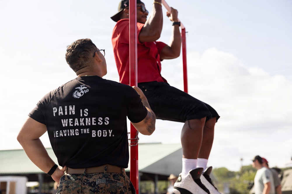 A U.S. Marine Corps instructor or recruiter, identified by the Eagle Globe and Anchor emblem and 'PAIN IS WEAKNESS LEAVING THE BODY' text on their black t-shirt and MARPAT camouflage pants with name tape 'MUNOZ', observes a person performing pull-ups on a red metal bar structure. The setting appears to be an outdoor fitness or recruitment event. The observer is monitoring the exercise performance of the individual on the bar.