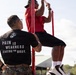 A U.S. Marine Corps instructor or recruiter, identified by the Eagle Globe and Anchor emblem and 'PAIN IS WEAKNESS LEAVING THE BODY' text on their black t-shirt and MARPAT camouflage pants with name tape 'MUNOZ', observes a person performing pull-ups on a red metal bar structure. The setting appears to be an outdoor fitness or recruitment event. The observer is monitoring the exercise performance of the individual on the bar.