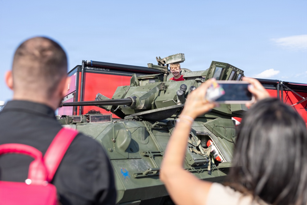 A young child in a red shirt is sitting in the open hatch of a military armored vehicle, smiling broadly. Two adults in the foreground — one carrying a pink bag, another holding a smartphone — are photographing the child. The setting appears to be a public military display or air show event.