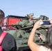 A young child in a red shirt is sitting in the open hatch of a military armored vehicle, smiling broadly. Two adults in the foreground — one carrying a pink bag, another holding a smartphone — are photographing the child. The setting appears to be a public military display or air show event.