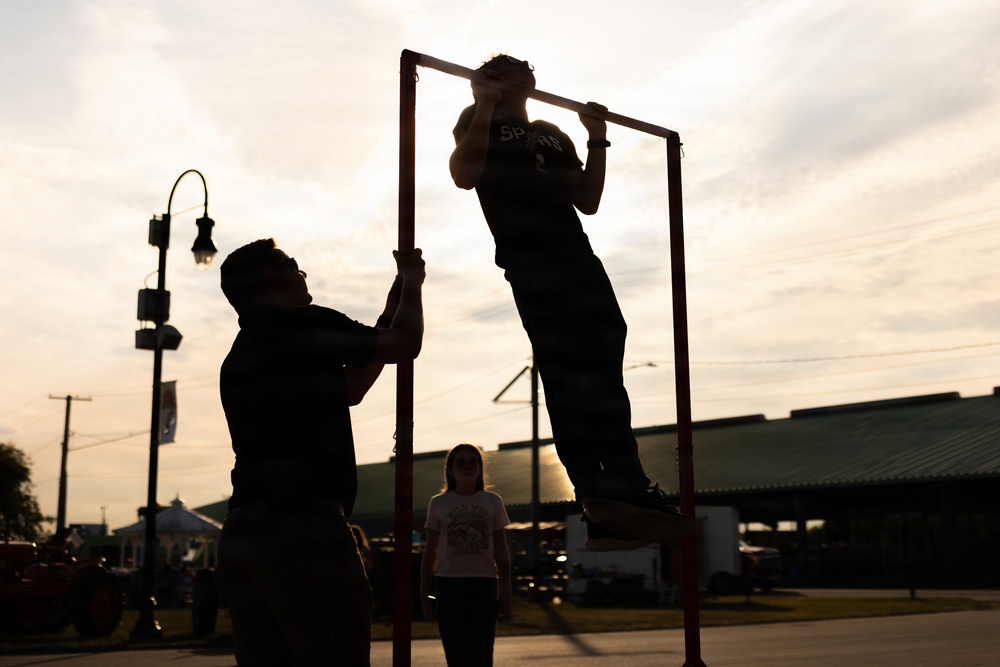A backlit silhouette scene captured at dusk or dawn shows two individuals interacting with a freestanding pull-up bar structure outdoors. One person is performing a pull-up or being lifted on the bar while another steadies the frame. A third person, appearing to be a child or young person, stands watching in the background.
