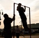A backlit silhouette scene captured at dusk or dawn shows two individuals interacting with a freestanding pull-up bar structure outdoors. One person is performing a pull-up or being lifted on the bar while another steadies the frame. A third person, appearing to be a child or young person, stands watching in the background.