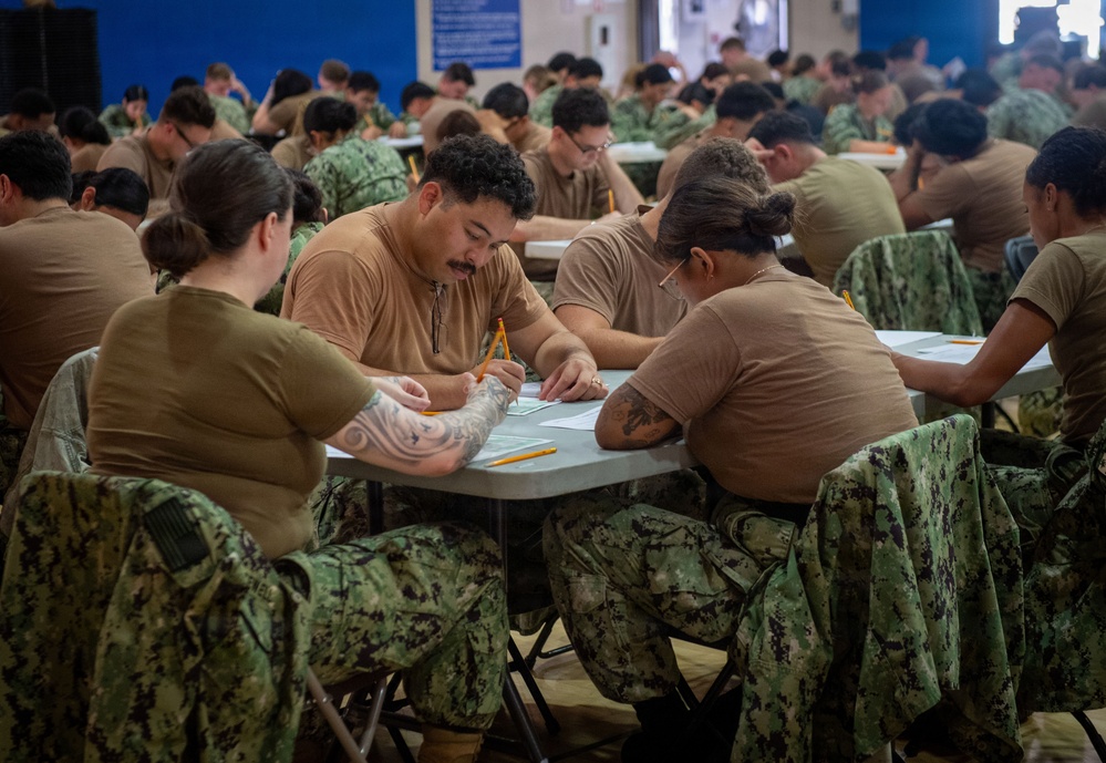 A large group of military personnel, appearing to be U.S. Navy sailors based on their NWU (Navy Working Uniform) camouflage pattern, are seated at tables in a large room taking what appears to be a written examination or completing paperwork. The individuals are wearing tan/coyote brown t-shirts with digital camouflage trousers. The setting resembles a gymnasium or large assembly hall configured for a mass testing event.