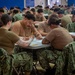 A large group of military personnel, appearing to be U.S. Navy sailors based on their NWU (Navy Working Uniform) camouflage pattern, are seated at tables in a large room taking what appears to be a written examination or completing paperwork. The individuals are wearing tan/coyote brown t-shirts with digital camouflage trousers. The setting resembles a gymnasium or large assembly hall configured for a mass testing event.