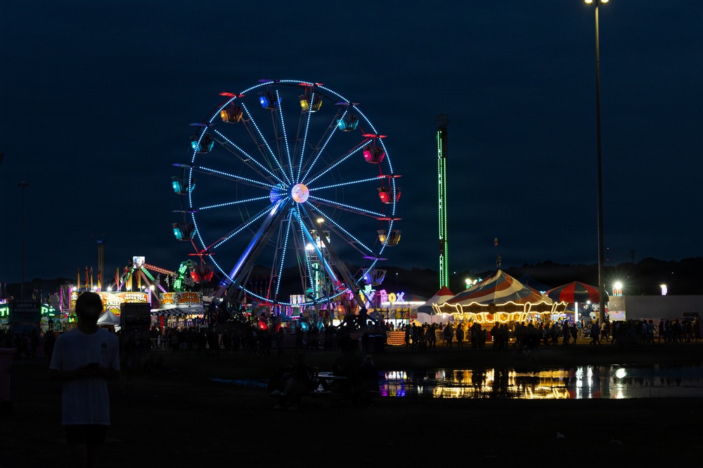A nighttime photograph of a county or state fair featuring a large illuminated Ferris wheel as the central focal point, surrounded by various carnival rides, tents, and crowds of people. The scene is reflected in a puddle of water in the foreground, adding depth to the composition. A silhouetted figure stands in the lower left, observing the fairgrounds.