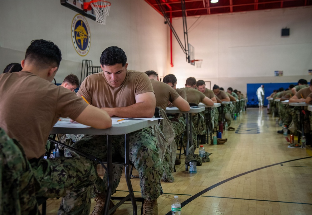 Military personnel in camouflage uniforms are seated at individual desks in a gymnasium, taking what appears to be a written examination or test. The setting is a military facility gymnasium, identifiable by the basketball hoop, hardwood floor, and an official seal on the wall reading 'Naval Medical Center San Diego.' Rows of service members extend deep into the background, all focused on their paperwork.