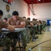 Military personnel in camouflage uniforms are seated at individual desks in a gymnasium, taking what appears to be a written examination or test. The setting is a military facility gymnasium, identifiable by the basketball hoop, hardwood floor, and an official seal on the wall reading 'Naval Medical Center San Diego.' Rows of service members extend deep into the background, all focused on their paperwork.