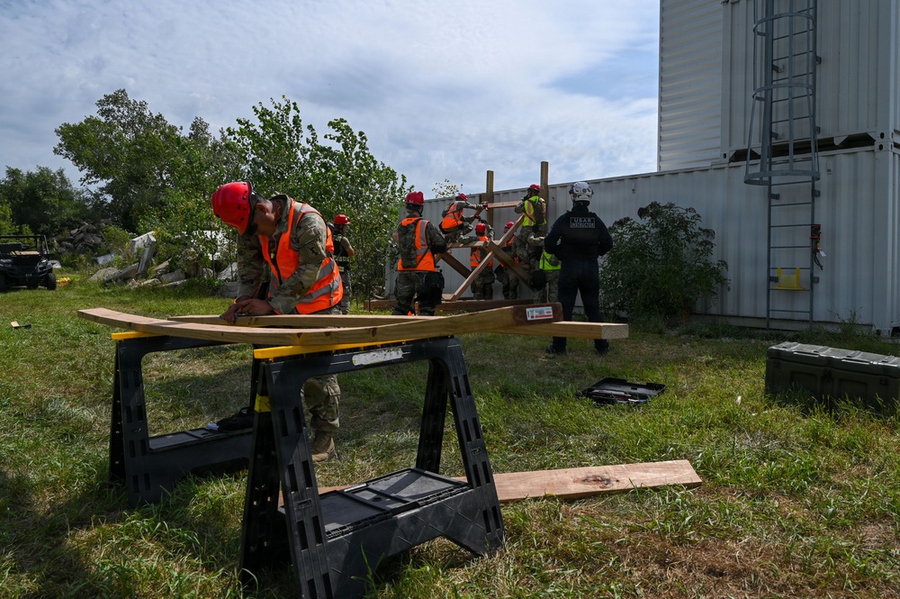 A group of personnel in military uniforms and orange safety vests with red hard hats are engaged in a structural/rescue training exercise outdoors. One individual in the foreground is working with lumber on a sawhorse workbench, while others in the background construct a wooden shoring or bracing structure near a large metal shipping container. An instructor or supervisor wearing a black 'USAR' (Urban Search and Rescue) uniform observes the group.