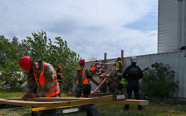 A group of personnel in military uniforms and orange safety vests with red hard hats are engaged in a structural/rescue training exercise outdoors. One individual in the foreground is working with lumber on a sawhorse workbench, while others in the background construct a wooden shoring or bracing structure near a large metal shipping container. An instructor or supervisor wearing a black 'USAR' (Urban Search and Rescue) uniform observes the group.