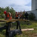 A group of personnel in military uniforms and orange safety vests with red hard hats are engaged in a structural/rescue training exercise outdoors. One individual in the foreground is working with lumber on a sawhorse workbench, while others in the background construct a wooden shoring or bracing structure near a large metal shipping container. An instructor or supervisor wearing a black 'USAR' (Urban Search and Rescue) uniform observes the group.
