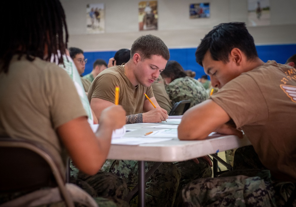 Military personnel in tan/brown t-shirts and camouflage pants are seated at folding tables in what appears to be a gymnasium or multipurpose room, taking a written examination or completing paperwork. The central figure is a young male with a forearm tattoo, holding a pencil and focused on his paper. Multiple service members are visible in the background engaged in the same activity.