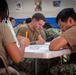 Military personnel in tan/brown t-shirts and camouflage pants are seated at folding tables in what appears to be a gymnasium or multipurpose room, taking a written examination or completing paperwork. The central figure is a young male with a forearm tattoo, holding a pencil and focused on his paper. Multiple service members are visible in the background engaged in the same activity.