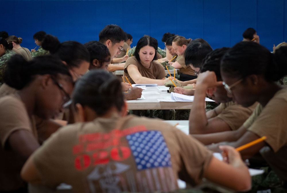 A large group of military personnel in uniform are seated at tables taking a written examination or completing paperwork. The individuals are wearing tan/khaki physical training shirts and camouflage uniforms consistent with U.S. military attire. The setting appears to be an indoor facility such as a gymnasium or large classroom with a blue wall visible in the background.