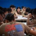 A large group of military personnel in uniform are seated at tables taking a written examination or completing paperwork. The individuals are wearing tan/khaki physical training shirts and camouflage uniforms consistent with U.S. military attire. The setting appears to be an indoor facility such as a gymnasium or large classroom with a blue wall visible in the background.
