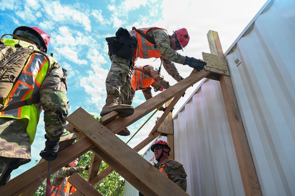 A group of military personnel in camouflage uniforms are working on a wooden structure, likely during a training exercise or disaster response operation. They are wearing high-visibility orange safety vests and red helmets while handling lumber on what appears to be a partially constructed or damaged wooden frame. The low-angle shot emphasizes the height and physical nature of the work being performed.